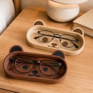 May include: Two oval-shaped bear-themed trays, one in light beige and one in dark brown, each featuring small rounded ears and a stylised bear face design on the base. A pair of black-rimmed glasses rests inside each tray.