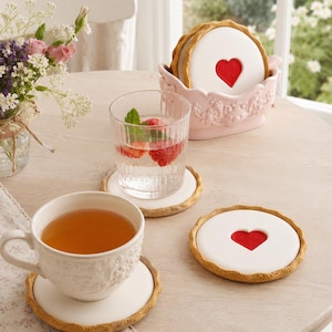 May include: A set of round drink coasters designed to look like shortbread biscuits with a white centre and a red heart cutout. The set includes a light pink decorative holder with embossed floral details, holding several stacked coasters.