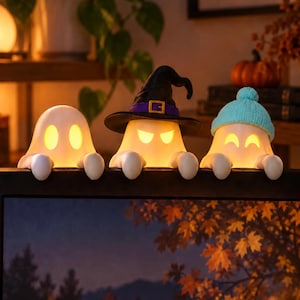 May include: Three illuminated ghost figures perched on a ledge. The left ghost is plain white, the centre ghost wears a black witch hat with a purple band, and the right ghost wears a light blue knit beanie. Each ghost has glowing yellow eyes.