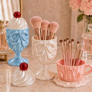 Peut inclure: Un récipient bleu clair avec couvercle et cerise, une coupe sur pied blanche contenant trois gros pinceaux de maquillage, et une tasse rose avec soucoupe contenant plusieurs petits pinceaux, ornés de nœuds et de perles en relief.