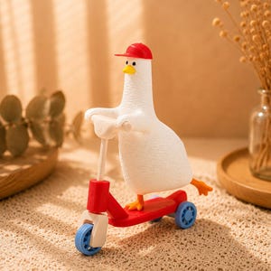 Puede incluir: Una figura de pato blanco con superficie texturizada que lleva una pequeña gorra de béisbol roja y un pico amarillo. El pato está colocado sobre un patinete rojo de tres ruedas con ruedas azul claro y manillar blanco, erguido.