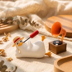 Op de afbeelding: Een wit eendenbeeldje met een strooien hoed met een rode band, liggend terwijl het een rode worst op een stokje vasthoudt. Naast de eend staat een bruine miniatuurgrammofoon met een oranje hoorn, een zwarte plaat en een witte knop.