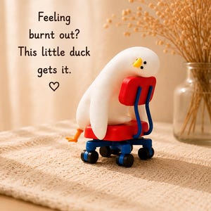 K&ouml;nnte beinhalten: Eine wei&szlig;e Entenfigur mit orangefarbenem Schnabel und F&uuml;&szlig;en, die auf einem rot-blauen B&uuml;rostuhl mit schwarzen Rollen zusammengesunken ist. Der Text lautet: Feeling burnt out? This little duck gets it. ♡