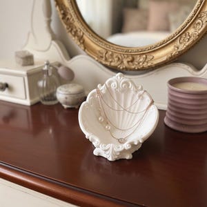 May include: A white, shell-shaped jewellery dish holds several necklaces with pearls and a small pendant. The dish sits on a dark wood surface, with a gold-framed mirror and a candle in the background. The jewellery dish is a decorative item.