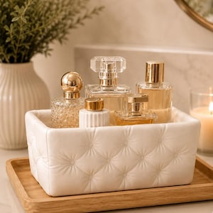 May include: A rectangular white storage bin with a tufted, button-detailed exterior texture. The bin is filled with five glass perfume bottles of varying shapes and sizes, featuring gold-toned caps and clear or amber-coloured liquid contents.