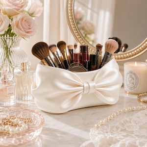 May include: A cream-coloured satin makeup brush holder featuring a large, centred bow design. The cylindrical container is filled with various makeup brushes with dark bristles and metallic handles, standing upright within the fabric-covered structure.