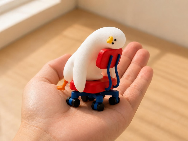 K&ouml;nnte beinhalten: Eine kleine wei&szlig;e Vogelfigur mit gelbem Schnabel und schwarzen Augen, die in einem roten Miniatur-B&uuml;rostuhl mit blauen Armlehnen und einem blauen Fu&szlig;kreuz mit f&uuml;nf schwarzen Rollen sitzt, wobei die orangefarbenen F&uuml;&szlig;e hinten sichtbar sind.