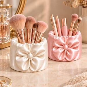 Peut inclure: Deux porte-pinceaux de maquillage cylindriques dotés d'un extérieur en tissu satiné froncé avec une grande fleur en tissu au centre. L'un est crème et contient des pinceaux, l'autre est rose clair et contient des stylos et des pinceaux.