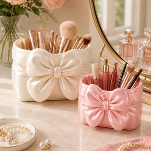 May include: Two oval-shaped makeup brush holders with a satin-like finish and a large decorative bow on the front. One holder is cream-coloured and the other is light pink. Both containers are filled with various makeup brushes and cosmetic tubes.