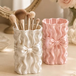 May include: Two cylindrical makeup brush holders with a glossy, ruffled texture and a prominent bow detail at the centre. One holder is pearlescent white and contains several makeup brushes with tan bristles, while the other is a solid pastel pink.