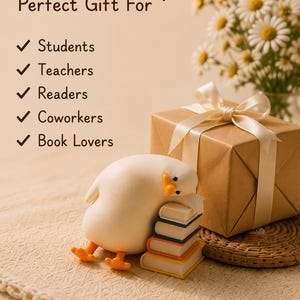 Puede incluir: Una figura de pato blanco descansando sobre una pila de tres libros peque&ntilde;os, junto a una caja de regalo marr&oacute;n atada con una cinta color crema. El texto dice: Perfect Gift For Students, Teachers, Readers, Coworkers, Book Lovers, con marcas.