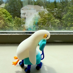 K&ouml;nnte beinhalten: Eine kleine wei&szlig;e Vogel-Figur mit langem Hals und orangefarbenen F&uuml;&szlig;en, die in einem leuchtend blauen B&uuml;rostuhl mit schwarzen Rollen sitzt. Der Vogel hat ein schlichtes Gesicht mit zwei schwarzen Punkten als Augen und einem kleinen gelben Schnabel.