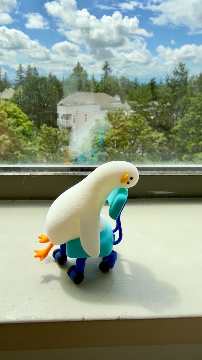 K&ouml;nnte beinhalten: Eine kleine wei&szlig;e Vogel-Figur mit langem Hals und orangefarbenen F&uuml;&szlig;en, die in einem leuchtend blauen B&uuml;rostuhl mit schwarzen Rollen sitzt. Der Vogel hat ein schlichtes Gesicht mit zwei schwarzen Punkten als Augen und einem kleinen gelben Schnabel.