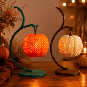Könnte beinhalten: Zwei dekorative Lampen mit kürbisförmigen Schirmen und Gittermuster. Eine Lampe hat einen geschwungenen türkisfarbenen Ständer mit einem orangefarbenen Kürbis, die andere einen braunen Ständer mit einem weißen Kürbis. Beide strahlen ein warmes Licht aus.