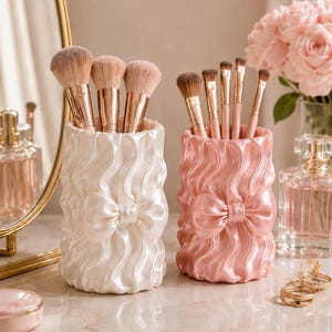 May include: Two cylindrical make-up brush holders with a vertical wavy, pleated texture and a central bow detail. One holder is cream-coloured and contains three large brushes, while the other is light pink and holds four smaller make-up brushes.