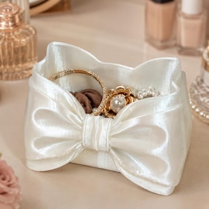 May include: A white satin fabric jewellery bowl featuring a prominent, structured bow design on the front. The interior of the bowl contains a gold-toned bracelet, a pearl-accented floral brooch, and a dark brown hair accessory.