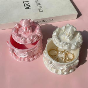 Puede incluir: Dos cajas redondas para joyas, una rosa claro y otra blanca, cada una con un oso esculpido en la tapa y detalles decorativos. La caja blanca está abierta, revelando anillos dorados y pendientes de perlas guardados en su interior.