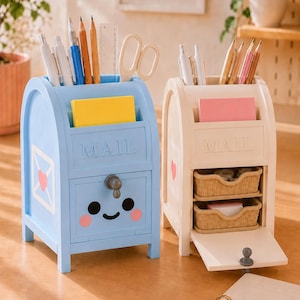 May include: Two mailbox-shaped desk organizers, one light blue and one cream, each featuring a top slot for mail, a front compartment, and a small drawer. The blue organizer has a smiling face, while the cream one has two small woven baskets.