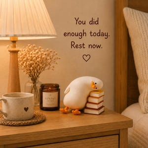Puede incluir: Una figura de pato blanco apoyando la cabeza sobre una pila de cuatro libros peque&ntilde;os, junto a una vela marr&oacute;n, una taza moteada con un coraz&oacute;n y un florero con flores secas. La pared muestra el texto: You did enough today. Rest now. con un coraz&oacute;n.