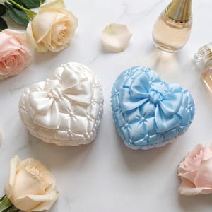 Pode incluir: Duas caixas de cetim em forma de coração, uma branca e outra azul clara, cada uma com um laço no topo. As caixas são rodeadas por rosas e frascos de perfume numa superfície branca.