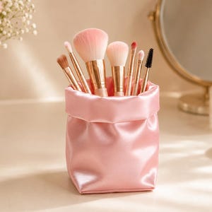 Peut inclure: Une pochette en satin rose doux remplie d'une collection de pinceaux de maquillage avec des viroles en or rose et des poils clairs. La pochette présente un bord supérieur replié et une finition lisse et réfléchissante, contenant divers pinceaux.