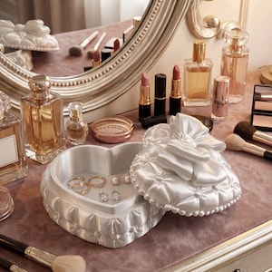 Könnte beinhalten: Eine Schminktisch-Szene mit einer herzförmigen Schmuckschatulle, Lippenstiften, Parfümflaschen und Make-up-Pinseln. Die weiße Schmuckschatulle enthält Ringe und Ohrringe. Ein großer, verzierter Spiegel spiegelt die Gegenstände wider.