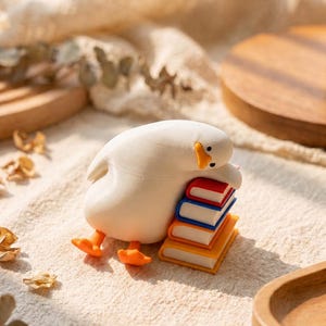 Puede incluir: Una peque&ntilde;a figura de pato blanco con patas naranjas y pico amarillo, inclinada sobre una pila de tres libros en miniatura. Los libros son de color rojo, azul y naranja, y el pato est&aacute; colocado como si estuviera apoyando su cabeza encima.