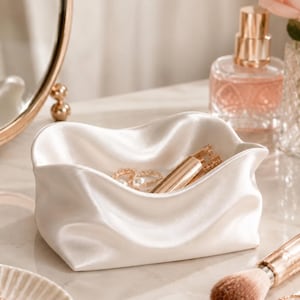 May include: A small, rectangular, off-white satin fabric tray with soft, gathered, and draped sides. The interior of the tray holds a gold-toned metal lipstick tube and a delicate gold ring with a small white pearl accent.
