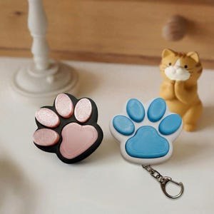 Może przedstawiać: Dwa breloczki do kluczy w kształcie łapy. Jeden jest czarny z różowymi podkładkami, drugi biały z niebieskimi podkładkami. W tle mała pomarańczowa figurka kota. Breloczki mają srebrny metalowy pierścień.