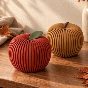May include: Two decorative apple-shaped objects with a ribbed, slatted design. One apple is deep red with a solid green leaf, and the other is a muted mustard yellow with a brown stem. Both items feature a rounded, segmented construction.