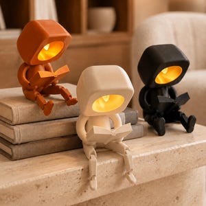 May include: Three articulated robot-shaped lamps in orange, cream, and black. Each figure has a large square head with a glowing yellow interior light and is depicted in a seated position while holding an open book in its hands.