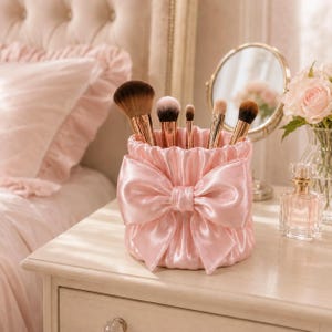 Puede incluir: Un soporte cilíndrico para brochas de maquillaje hecho de tela de satén rosa claro fruncido, con un gran lazo decorativo de satén en la parte frontal. El soporte contiene cinco brochas de maquillaje variadas con cerdas marrones y mangos dorados.