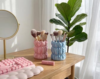 May include: Two decorative makeup brush holders, one pink and one blue, sit on a wooden table. Each holder is filled with makeup brushes. A pink keyboard and a lipstick are also on the table. A gold-framed mirror and a green plant are in the background.