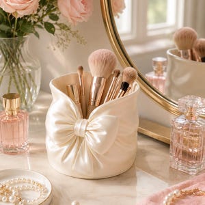 Puede incluir: Un porta brochas de maquillaje cilíndrico de color crema con un gran lazo de satén en la parte frontal. El soporte contiene varias brochas de maquillaje con mangos metálicos, situado junto a frascos de perfume y joyas de perlas.