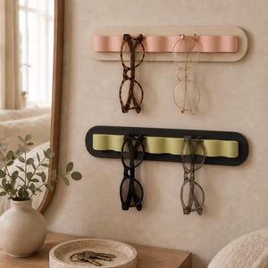 Peut inclure: Deux organisateurs muraux horizontaux dotés de crêtes ondulées en relief. L'organisateur supérieur est beige avec un insert rose, supportant deux paires de lunettes. L'inférieur est noir avec un insert vert clair, supportant deux paires de lunettes.