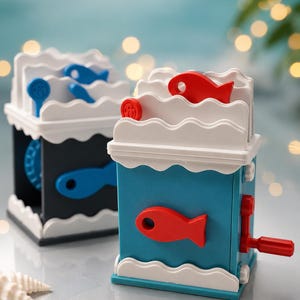 Puede incluir: Dos juguetes mecánicos cuadrados con diseño de olas en la parte superior e inferior. Uno es azul con manivela y peces rojos, el otro es negro con manivela y peces azules. Ambos cuentan con detalles decorativos blancos en forma de onda.