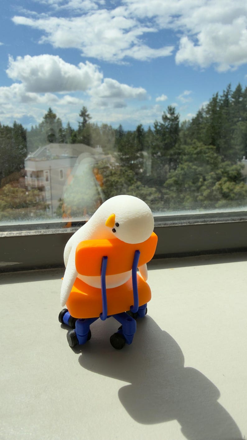 K&ouml;nnte beinhalten: Eine kleine wei&szlig;e Figur mit rundem Kopf und gelbem Schnabel, die auf einem orangefarbenen Miniatur-B&uuml;rostuhl mit blauen St&uuml;tzen und schwarzen Rollen sitzt. Der Stuhl hat eine R&uuml;ckenlehne und eine Sitzfl&auml;che, &uuml;ber die sich die Figur beugt.