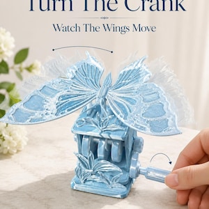 Puede incluir: Una escultura de mariposa mecánica de color azul claro con alas translúcidas montada sobre una base con una manivela manual. El texto Turn The Crank y Watch The Wings Move aparece sobre el objeto, que presenta intrincados patrones en las alas.
