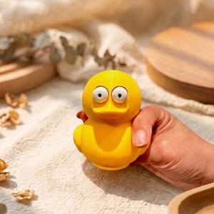 Pode incluir: Um brinquedo de pato de borracha amarelo brilhante com dois olhos brancos grandes e arregalados com pupilas pretas na frente da cabeça. O pato tem um corpo liso e arredondado e um bico plano, com um design simples e estilo de desenho animado.