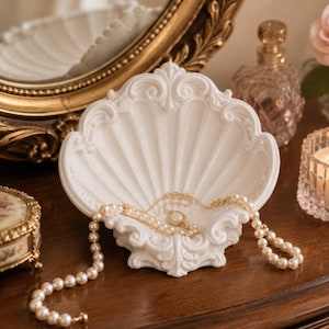 Könnte beinhalten: Eine weiße, muschelförmige Ablageschale aus Keramik mit kunstvollen, erhabenen Verzierungen an den Rändern. Eine zarte, goldfarbene Halskette mit einem kleinen Anhänger und eine Kette aus cremefarbenen Kunstperlen liegen in der Schale.