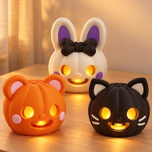 May include: Three illuminated pumpkin-shaped lamps featuring animal designs. One is orange with bear ears, one is white with rabbit ears and a black bow, and one is black with cat ears. Each lamp has glowing yellow eyes and a smiling mouth.