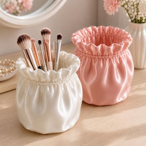 May include: Two round, drawstring-style pouches made of smooth, shiny fabric. One pouch is white and holds several makeup brushes with metallic handles and soft bristles. The second pouch is light pink and sits empty next to the white one.