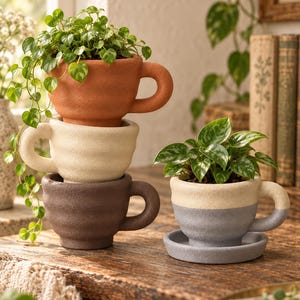 May include: A stack of three textured ceramic mug-shaped planters in terracotta, cream, and dark brown, each holding a green plant. Beside them sits a fourth mug-shaped planter in cream and grey, resting on a matching saucer with a green plant.