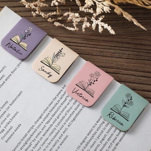 Puede incluir: Cuatro marcadores de libros de cuero personalizados en colores pastel: lavanda, beige, rosa y verde menta. Cada uno presenta un dise&ntilde;o floral, una ilustraci&oacute;n de libro abierto y un nombre: Natalie, Sandy, Victoria y Rebecca. Los marcadores est&aacute;n en una p&aacute;gina blanca.