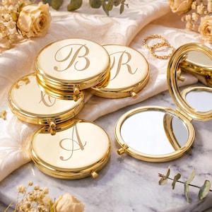 Puede incluir: Una colección de espejos de bolsillo redondos de metal dorado con superficies pulidas. Varios espejos están cerrados y presentan letras cursivas grabadas, incluidas A, B y R. Un espejo está abierto, revelando un interior de cristal circular.