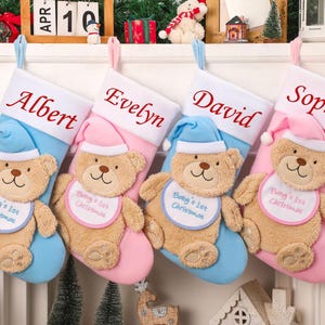 May include: Four Christmas stockings, each with a name embroidered in red: Albert, Evelyn, David, and Sophia. Each stocking has a teddy bear with a bib that says "Baby's 1st Christmas". The stockings are blue and pink.