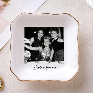 May include: White ceramic trinket dish with a gold rim, featuring a black and white photo of three young women taking a selfie. The words "Bestie forever" are printed below the photo. A ring is visible in the lower left corner.