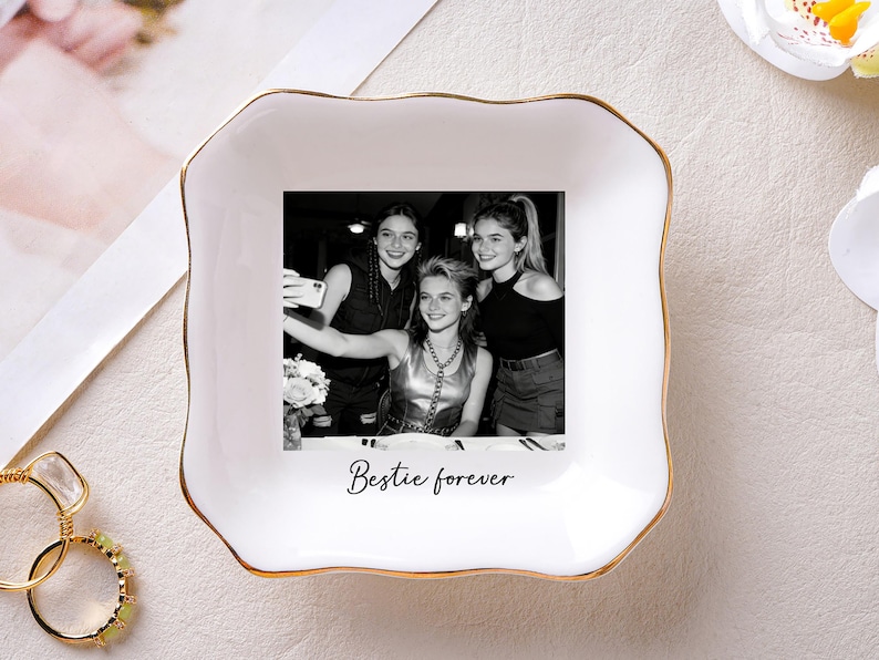 May include: White ceramic trinket dish with a gold rim, featuring a black and white photo of three young women taking a selfie. The words "Bestie forever" are printed below the photo. A ring is visible in the lower left corner.