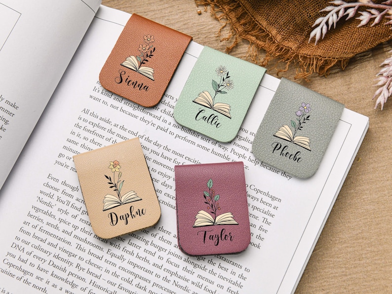 Puede incluir: Cinco marcadores de cuero con dise&ntilde;os florales y nombres: Sienna, Callie, Phoebe, Daphne y Taylor. Cada marcador es de un color diferente, incluyendo marr&oacute;n, verde, gris, beige y burdeos. Est&aacute;n colocados en un libro abierto.