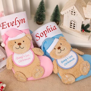 May include: Two personalized Christmas stockings, one pink and one blue, each with a teddy bear design. The pink stocking is labeled "Evelyn" and the blue stocking is labeled "Sophia". Each bear wears a Santa hat and bib.